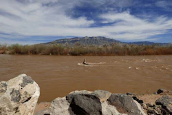 Muddy river flowing through desert landscape with mountains and bare trees under blue sky