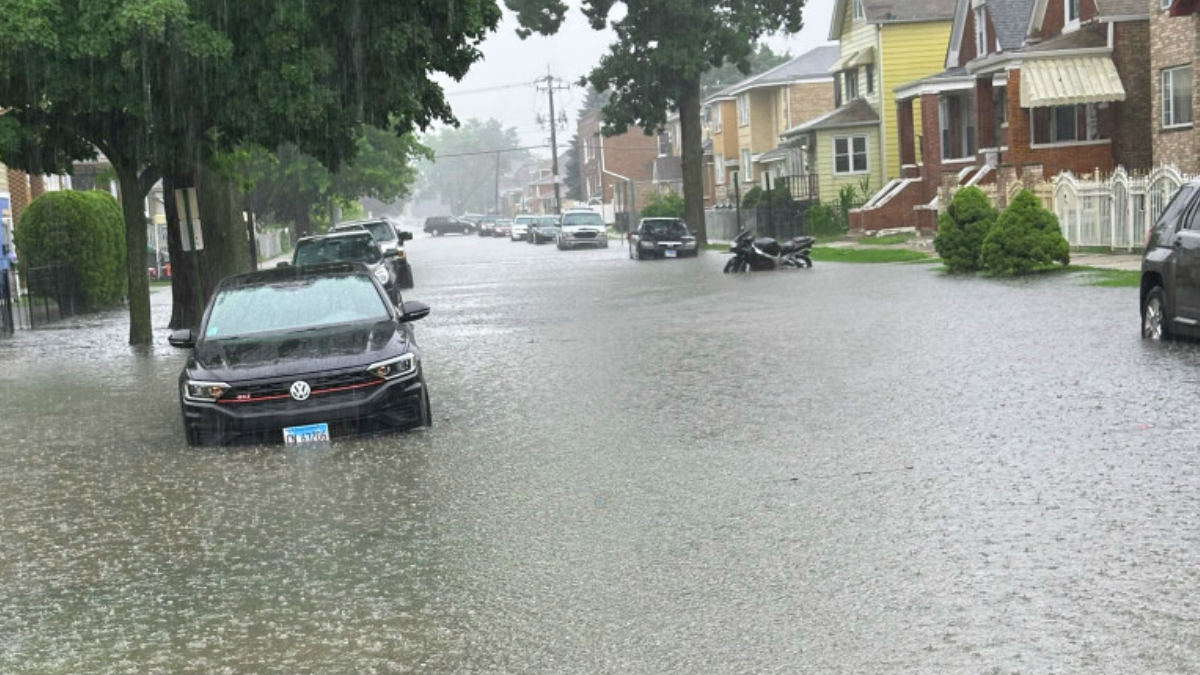 Residential street flooded with water, parked vehicles partially submerged, brick homes and trees visible in heavy rain.