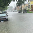 Residential street flooded with water, parked vehicles partially submerged, brick homes and trees visible in heavy rain.
