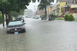 Residential street flooded with water, parked vehicles partially submerged, brick homes and trees visible in heavy rain.