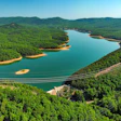 Aerial view of turquoise reservoir surrounded by dense green forest and mountains with a bridge crossing the water