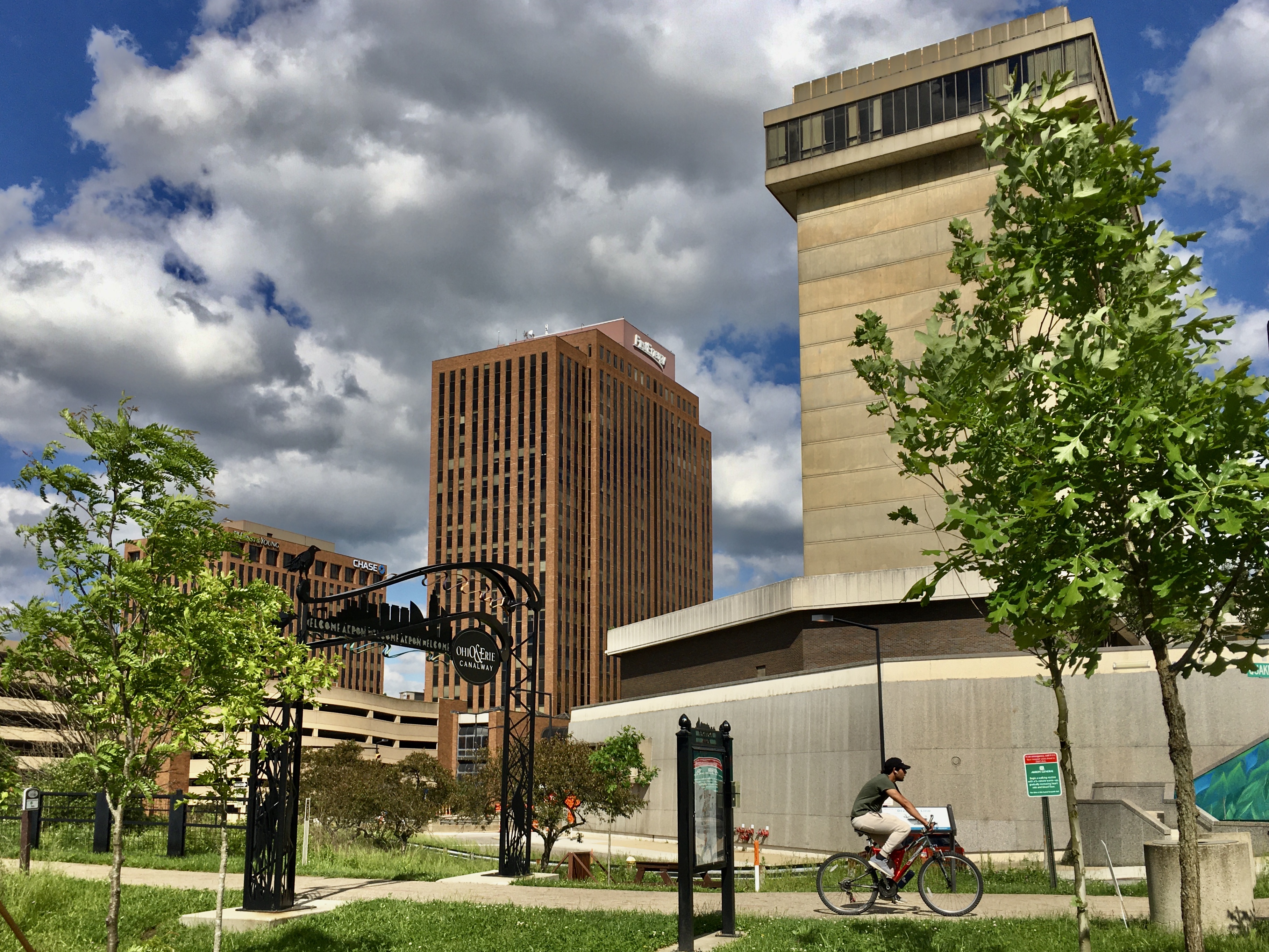 Urban park with modern architecture, featuring tall buildings, green trees, and cyclist on pathway under cloudy sky