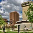 Urban park with modern architecture, featuring tall buildings, green trees, and cyclist on pathway under cloudy sky